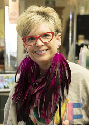 closeup of a woman with short blond hair, bright red glasses and funky magenta feathered earrings that hang down to her chest