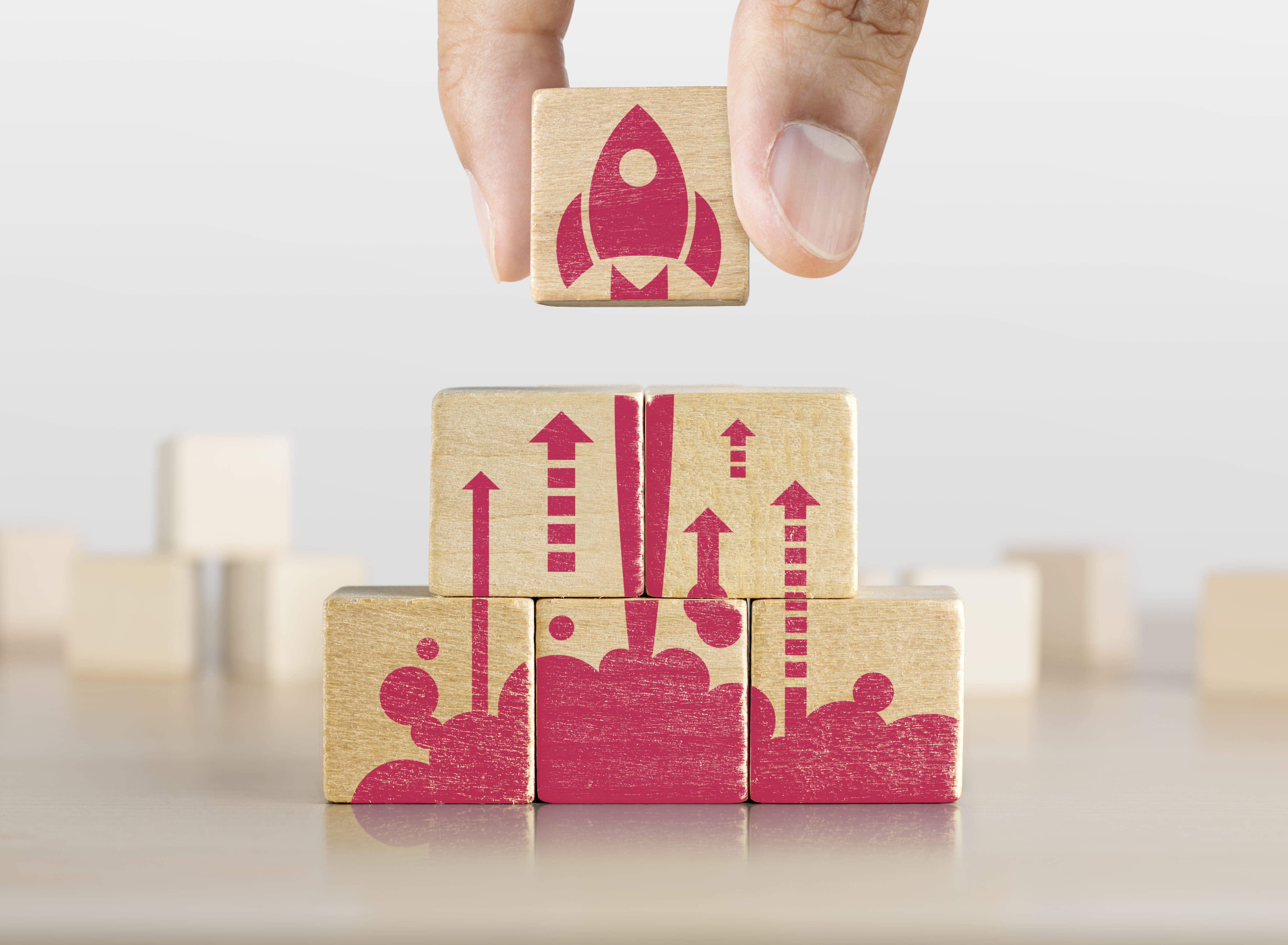 Wooden blocks stacked on a table. Painted on the blocks is an image of a rocket ship blasting off. A hand can be seen stacking the last block on top.