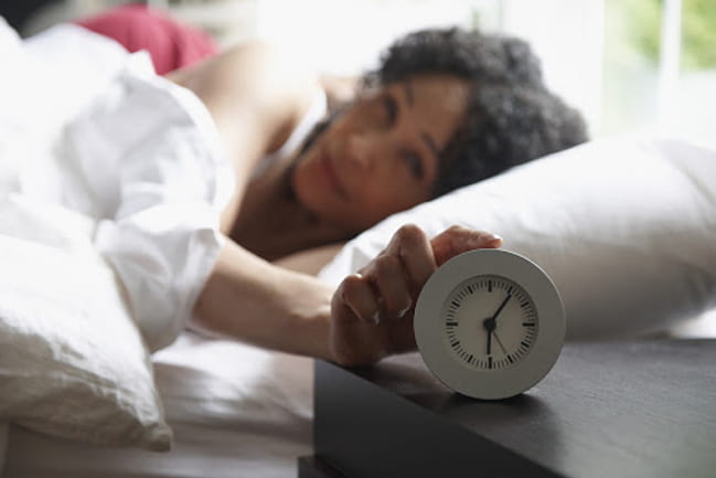 Person laying in bed, snoozing their alarm clock on the nightstand