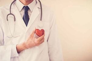 A doctor in white coat and a stethoscope around his neck holds a red heart shape in front of him. 