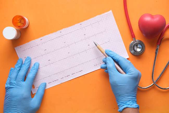 Image of rubber-gloved hands holding a pencil looking at a cardiology report with a stethescope, foam heart, and open pill bottle nearby.
