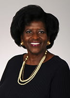 Aurelia Sands-Belle is wearing a black blouse and a pearl necklace and is smiling at the camera.