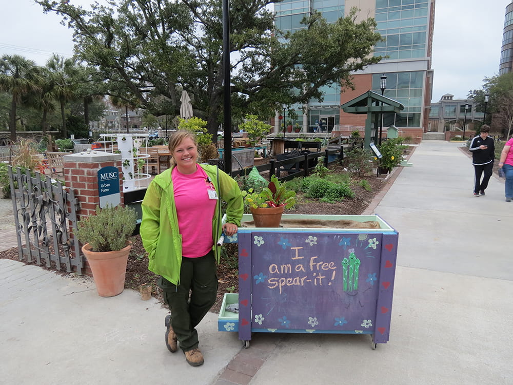 Carmen Ketron poses in front of the Urban Farm alongside a chalkboard that says "I am a free spear-it" with cartoon-y drawings of asparagus spears