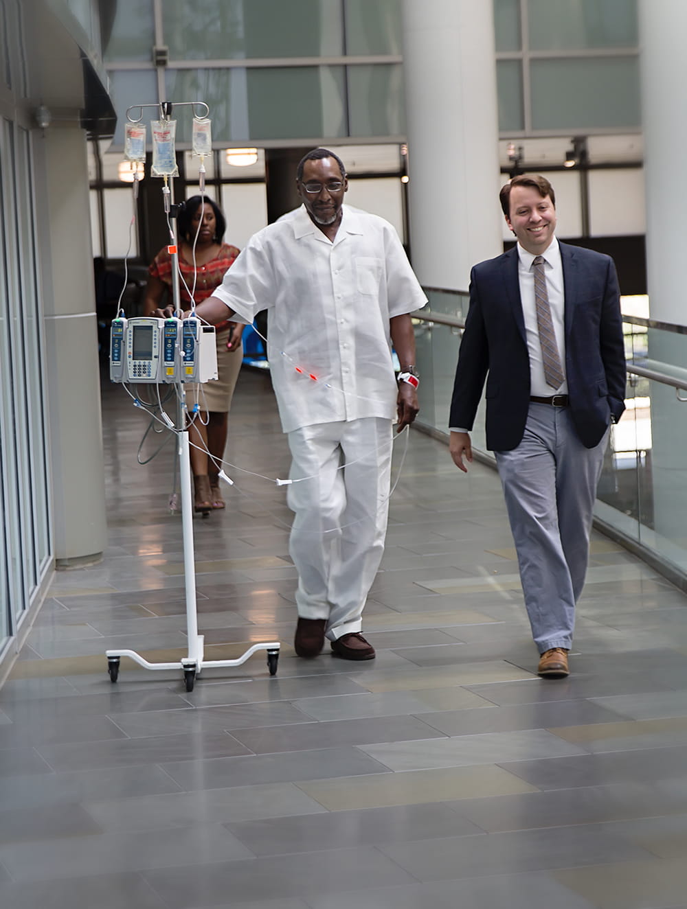 doctor in sports coat walks alongside groom dressed in white shirt and pants but attached to IV pole down the chrome and glass hallway in the Ashley River Tower at MUSC