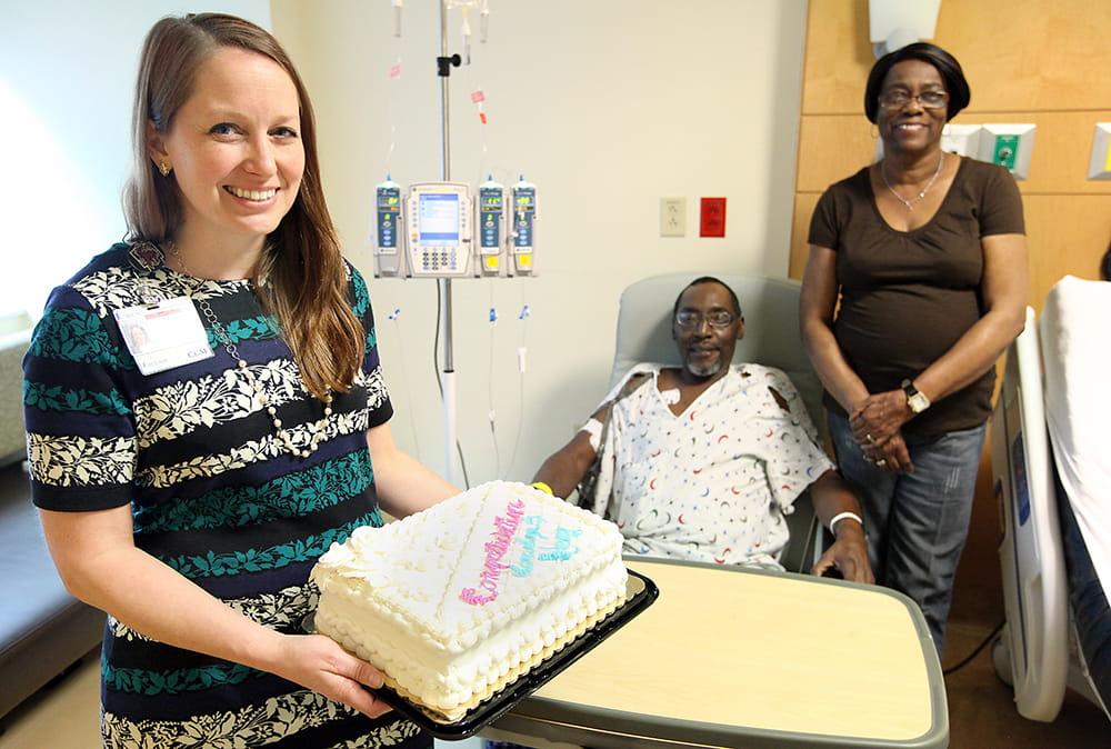 Nurse case manager Amanda Ghent displays the wedding cake while the bride and groom look on in the groom's hospital room