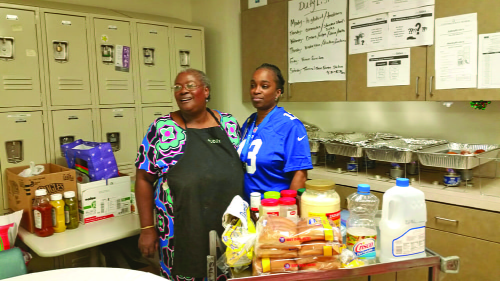 Two women stand in an employee break room surrounded by food items. 