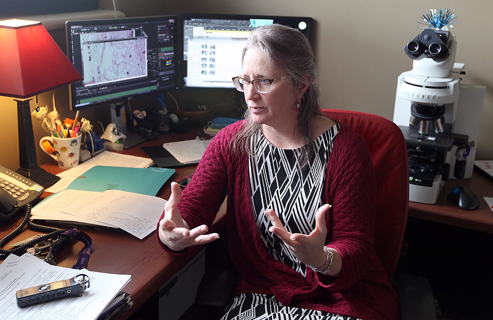 Kristi Helke sits at a desk covered with papers, a computer monitor and microscope and gestures as she speaks. 