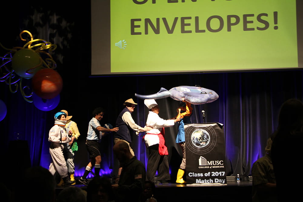 Deans dressed as an farmer, astronaut, soccer player, safari guide, chef and fisherman dance in a conga line on stage. 