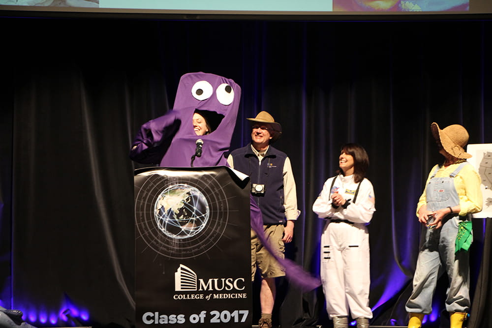 A woman in a purple google-eyed costume speaks at a podium. 