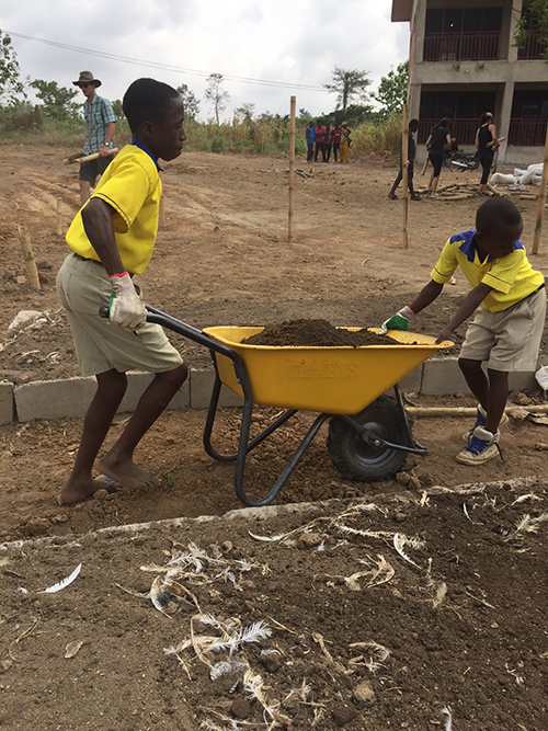 Two boys in school uniform navigate with a large wheelbarrow filled with dirt -- one pushing and one pulling