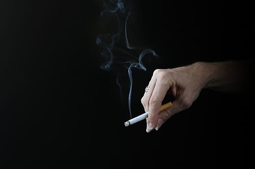 a hand holds a cigarette against a black background as smoke wafts into the air 