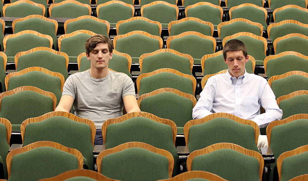 two students sit amid a theater-like auditorium of empty chairs with their eyes closed as they meditate