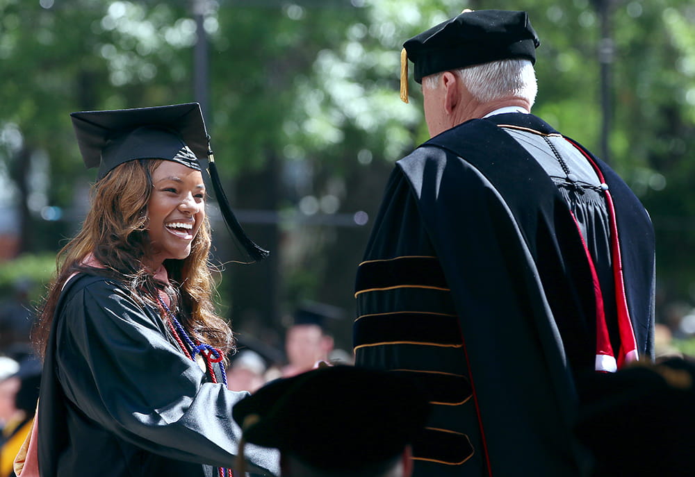 A smiling woman in cap and gown shakes hands with the university president 