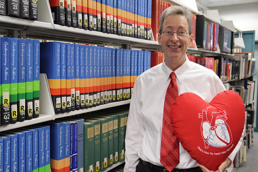 A man stands in front of a shelf of books at the library and holds a bright red pillow shaped liked a heart 