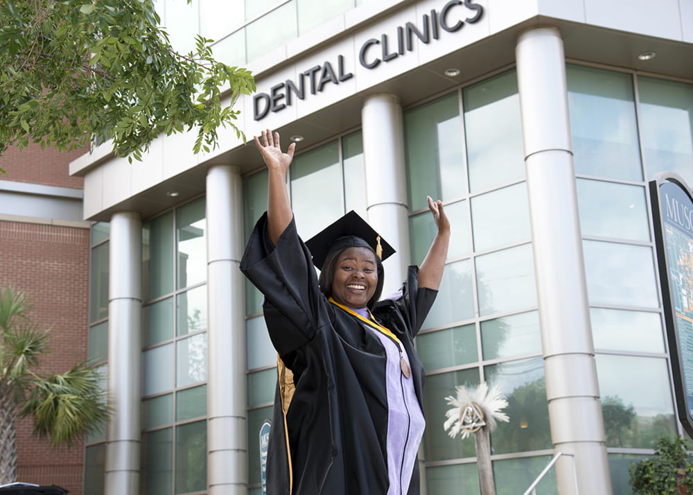 A woman in cap and gown holds her hands overhead in victory while standing in front of the dental clinic building 