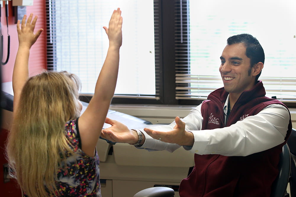 A seated doctor holds his arms out in front of his body while facing a young girl who has her arms raised overhead