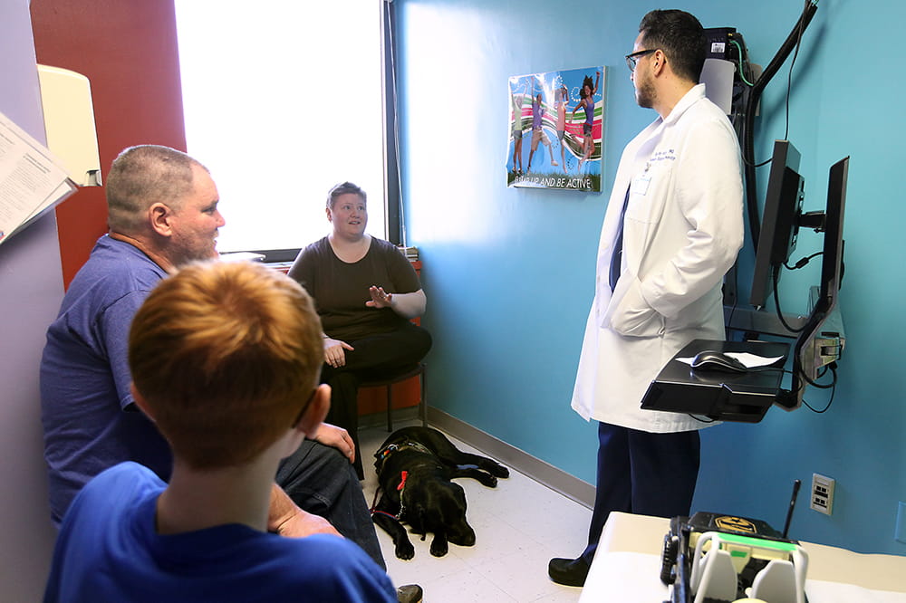 A family sits in an exam room and speaks to a doctor as the dog chills on the floor