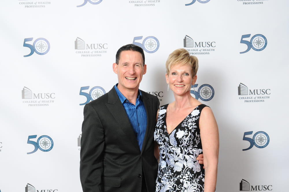 A couple poses in front of a backdrop with '50' and "MUSC College of Health Professions" displayed