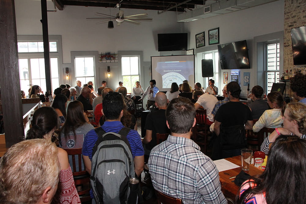a large crowd sits within a bar and listens to speakers standing in front of a screen