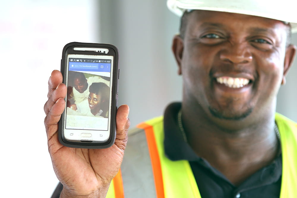 A worker in safety vest and hard hat holds up a phone showing a Facebook photo of himself, his wife and their newborn at MUSC years ago