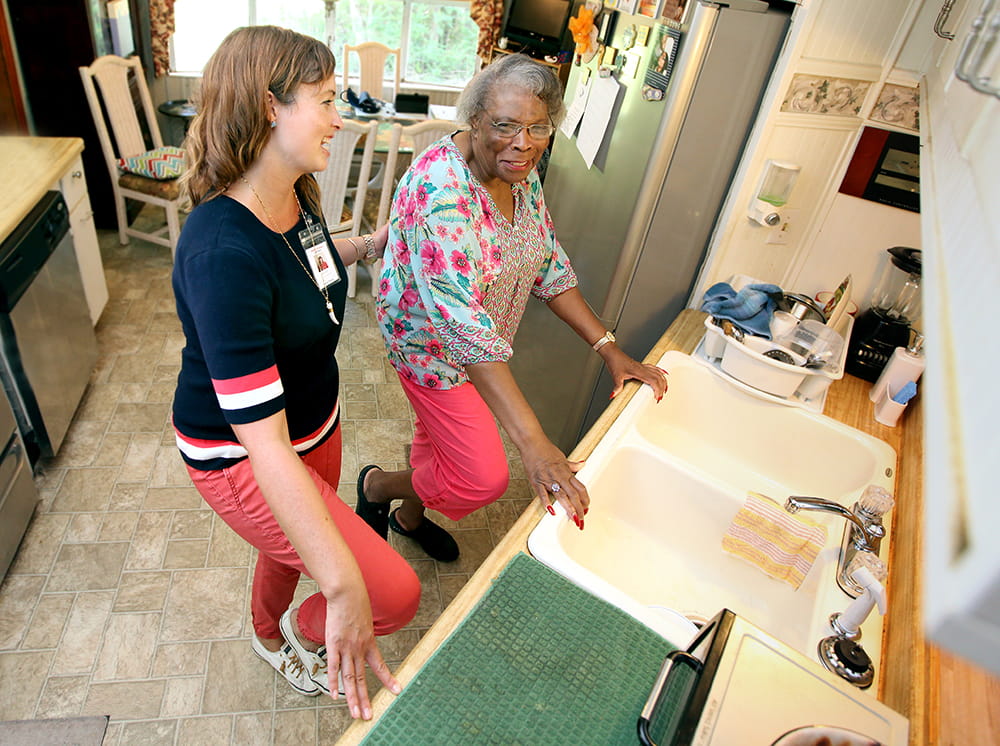 An older woman grasps the kitchen counter with both hands and lifts her right knee. Next to her a young woman has one steadying hand on the older woman's back and one hand on the counter as she lifts her knee to demonstrate