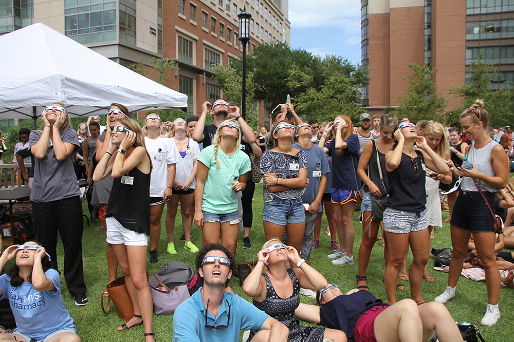 a group of people sitting and standing on a lawn look up at the sky with their eclipse glasses on