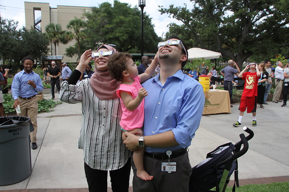 A toddler in her father's arms tugs at his strange eclipse glasses as he and his wife look up at the sky
