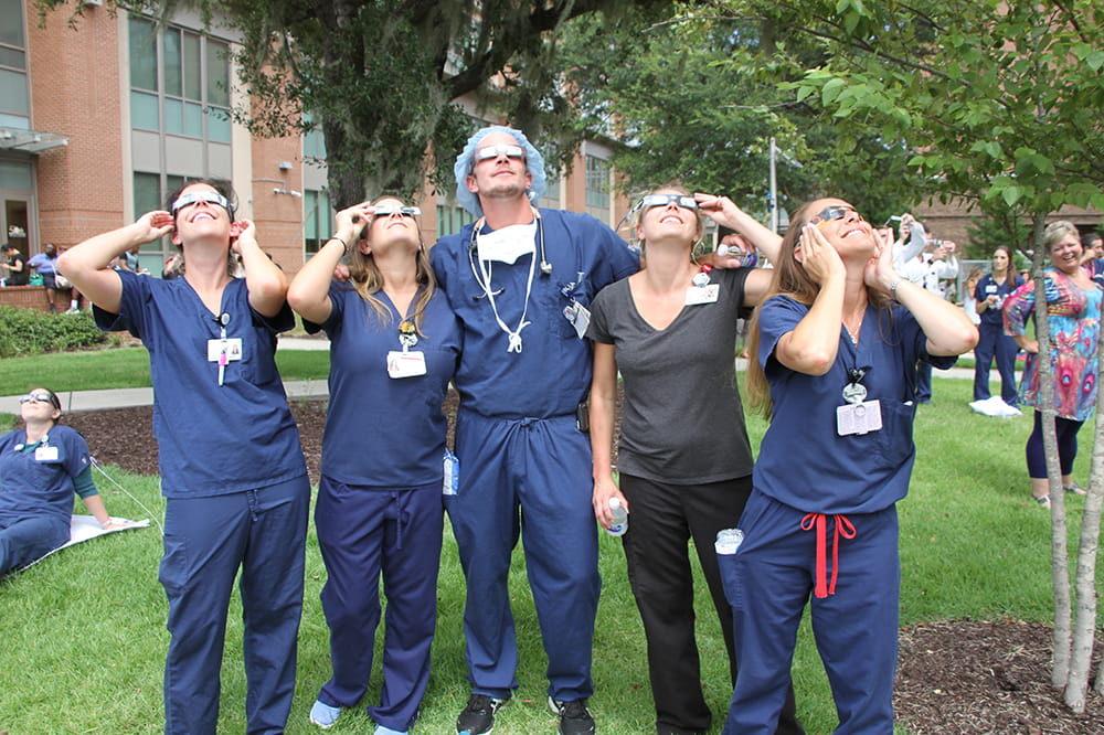 a group of five nurses, four in dark blue scrubs, look up at the sky while wearing eclipse glasses