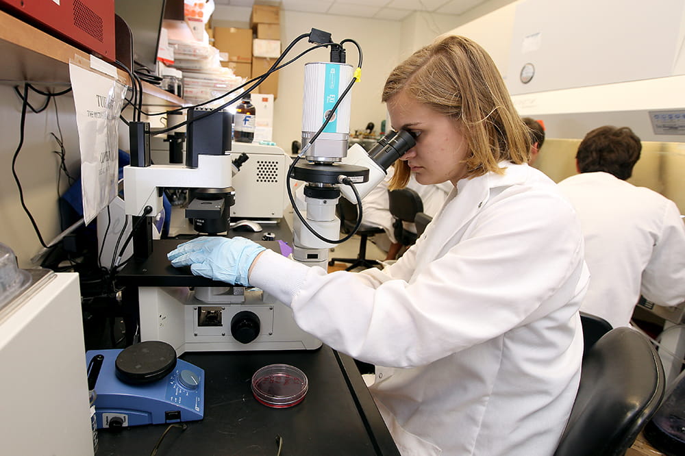 A young woman in white lab coat peers into a microscope