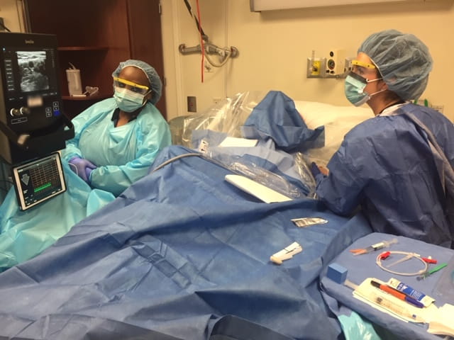 Two women in scrubs prepare for a bedside procedure