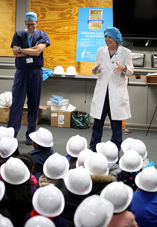 Dr. Robert Cina and PA Helen Kulseth stand in front of a group of small children wearing plastics hard hats and seated on the floor