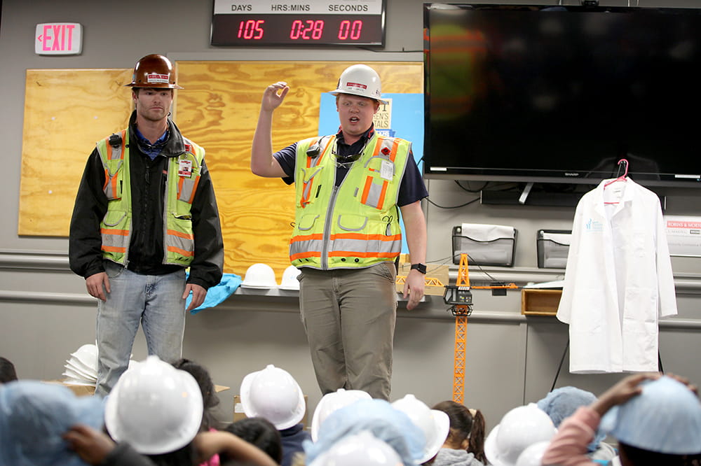 two construction workers in safety gear talk to a group of children seated on the floor 