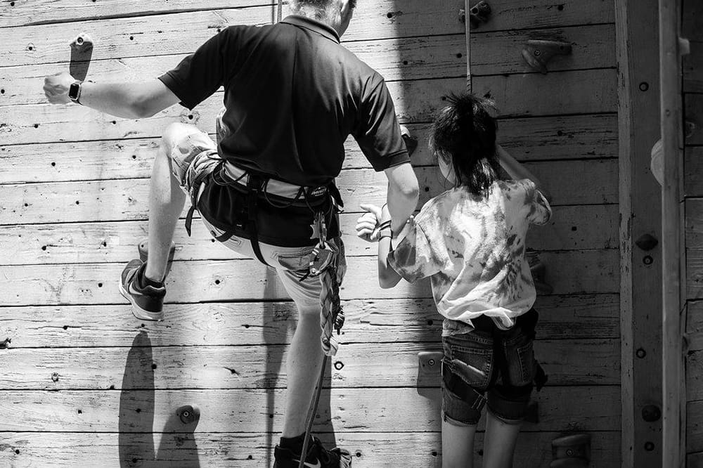 A child and a counselor begin climbing up a climbing wall