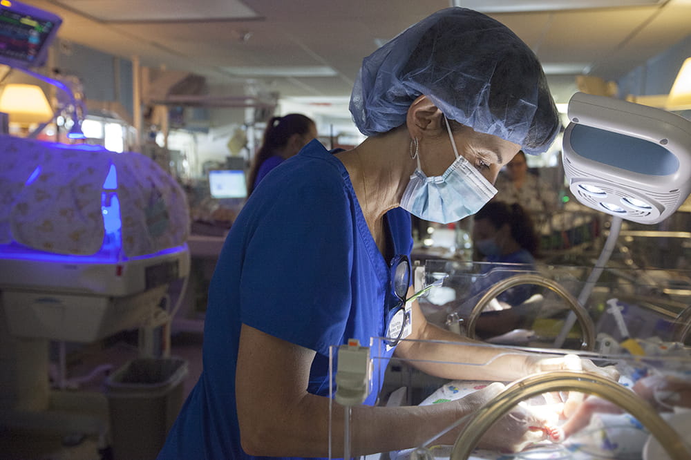 Wearing a mask and hair covering, Paulette Headden works with a tiny baby in the NICU