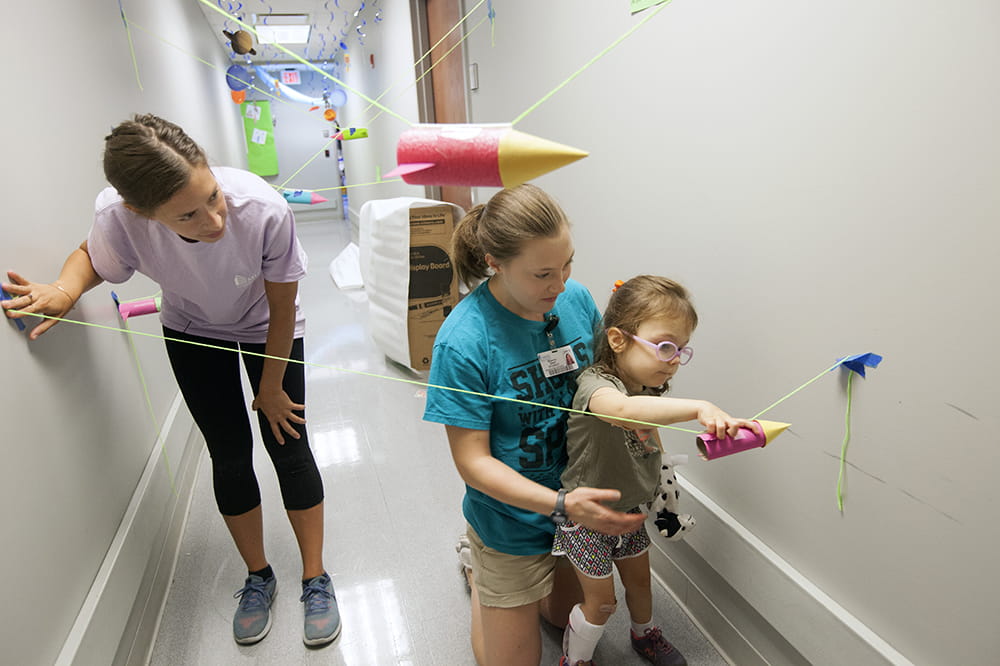 a little girl concentrates as she moves a cardboard tube along a string while a student helps her stand