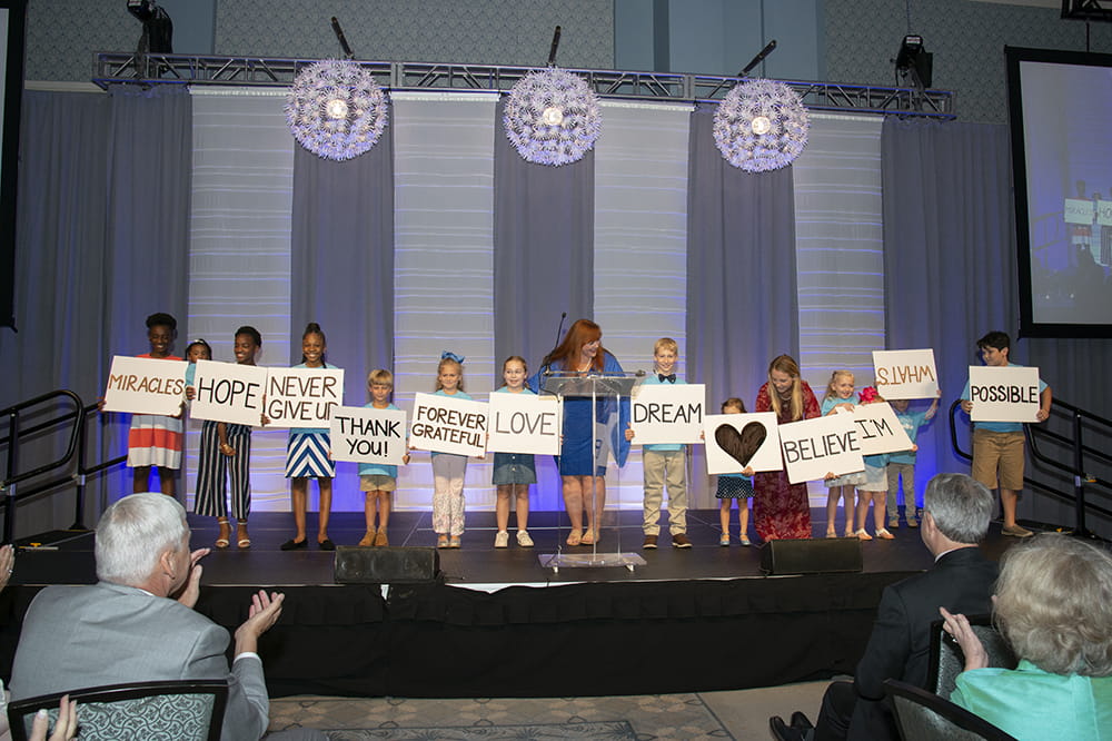 Current and former patients stand on a stage and hold up signs with messages like Hope, Love and Forever Grateful. 