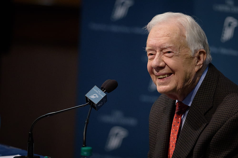 an elderly Jimmy Carter smiles while at a microphone