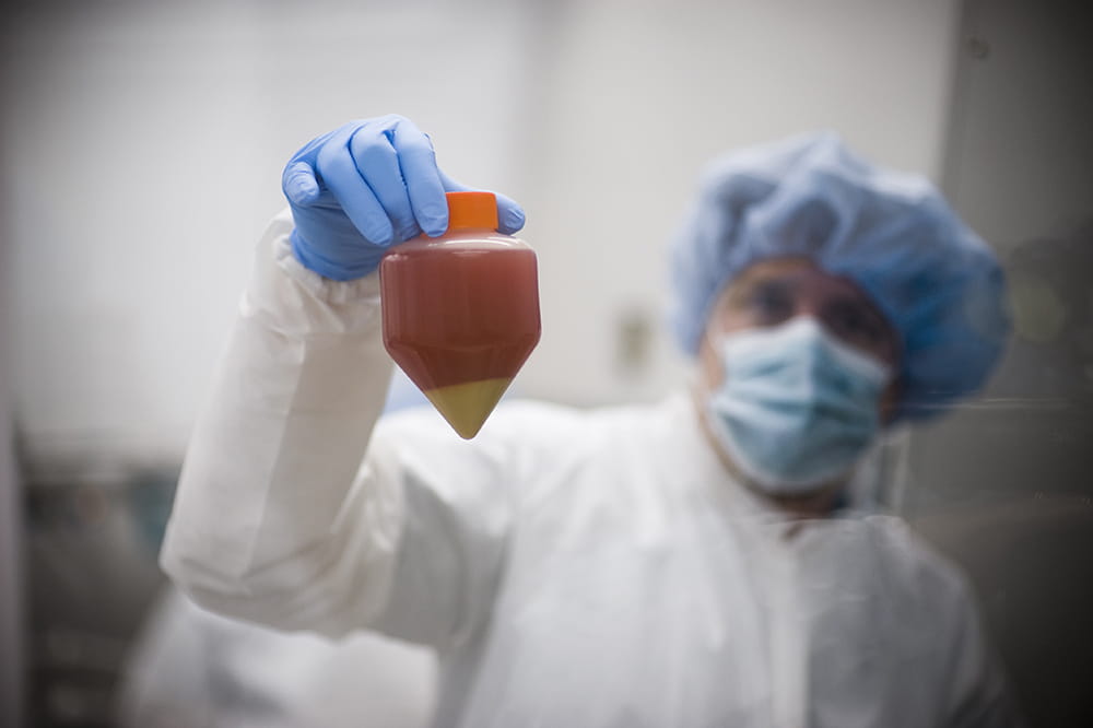 A worker wearing coverings over the hair, face, body and hands holds up a plastic container. Inside is a liquid that has separated into a reddish liquid above and a yellow liquid at the bottom. 