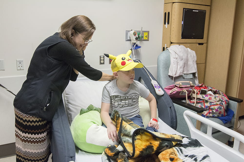 A doctor uses a stethoscope to listen to a young boy's breathing as he sits in his hospital bed wearing a Pokemon hat. 