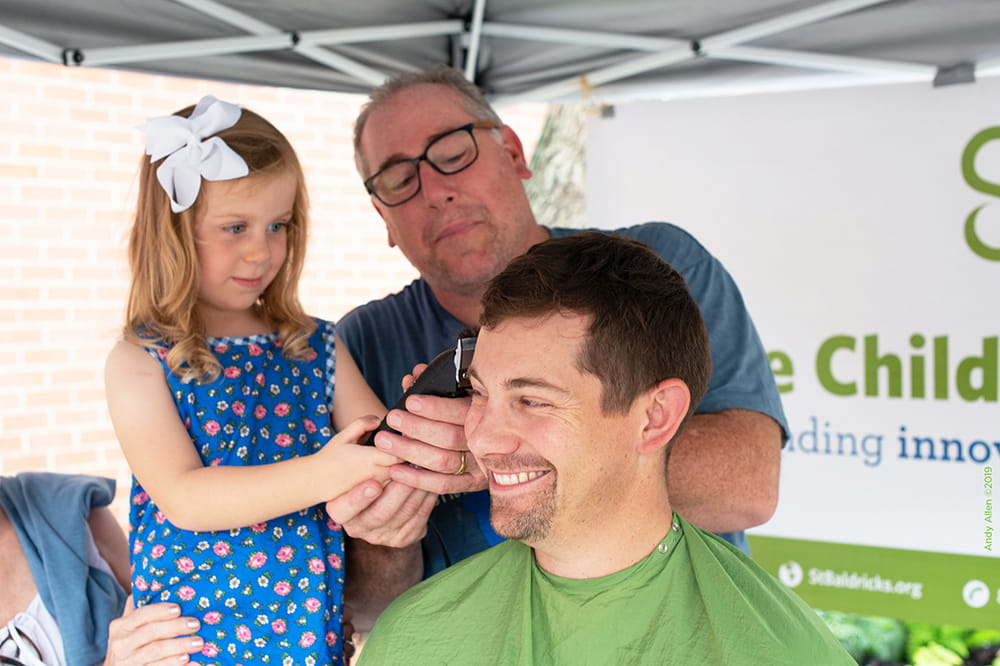 A man laughs as child and another man shave his head. 