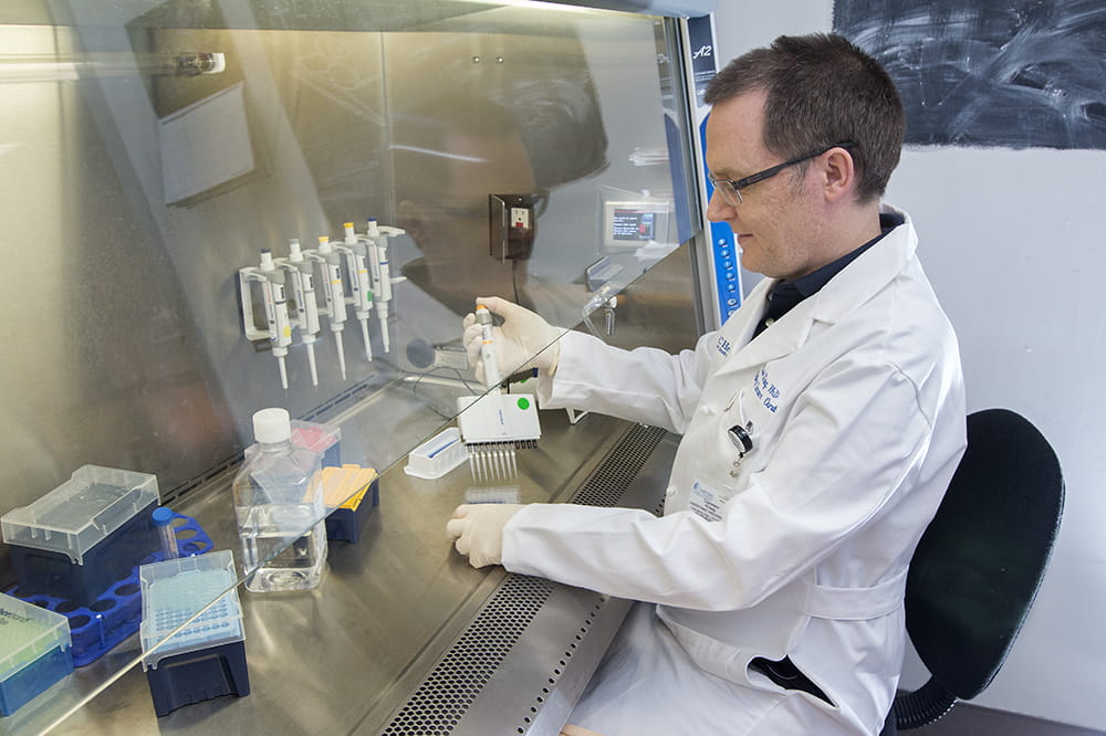 A man in a white lab coat sits at a table and pipettes samples behind a transparent shield