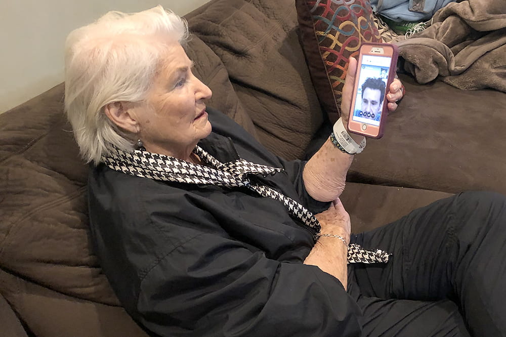 an older woman sits on a couch looking at a smartphone. Her doctor's face is visible on the screen