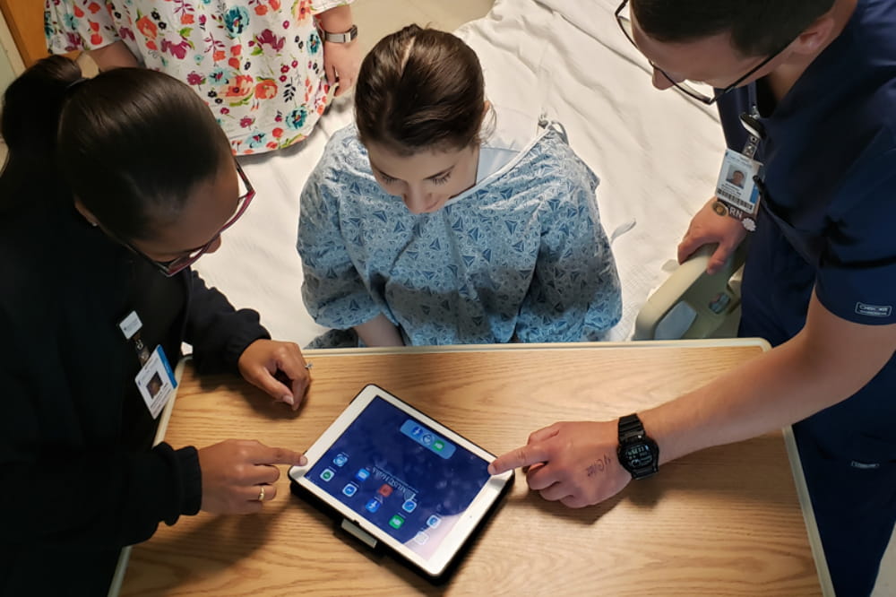 a woman in a hospital gown looks at an ipad as two health care workers show her how it works