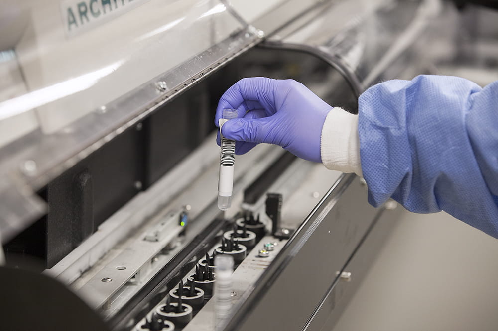 a closeup shot of a gloved hand placing a vial of blood into a giant machine