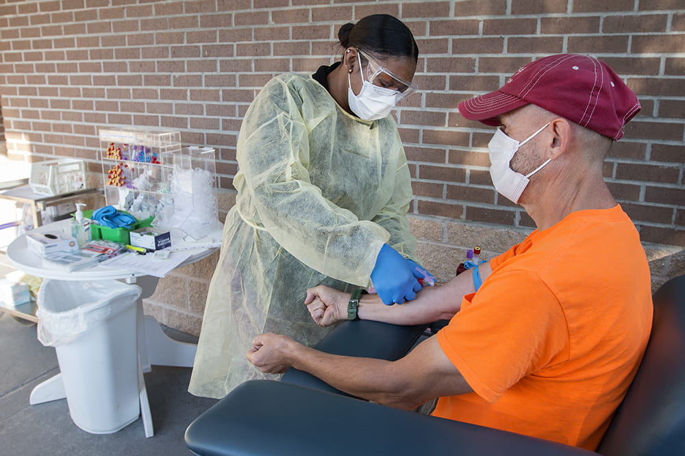 a masked, gloved and gowned woman preps the arm of a masked older man outside a brick building