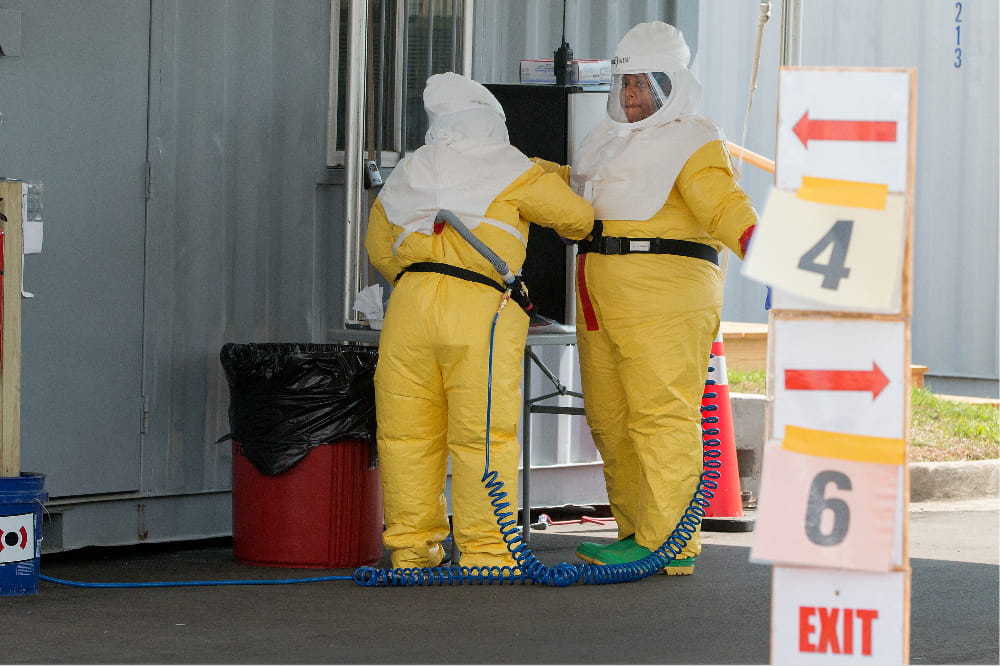 two women check each other's PPE. They are covered head to toe, wearing beekeeper-style hoods and baggy yellow jumpsuits 