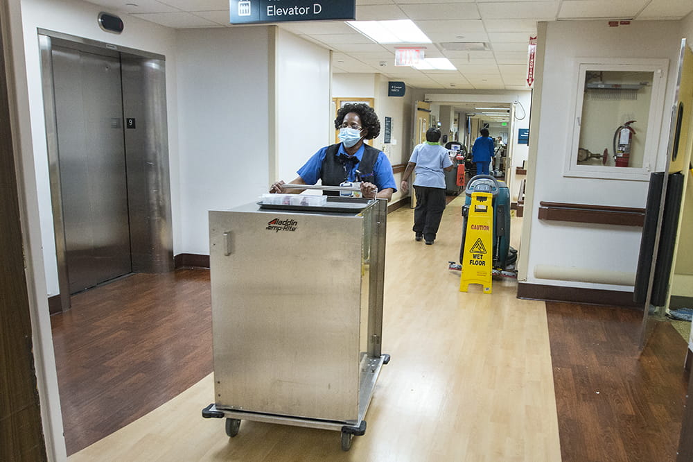 A woman pushes a large, enclosed stainless steel cart down a hospital hallway. The only other people in the hallway are other food service and environmental service employees.