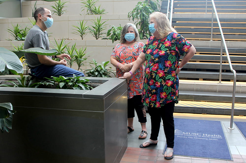 A man sits on the edge of an indoor planter chatting with two standing women