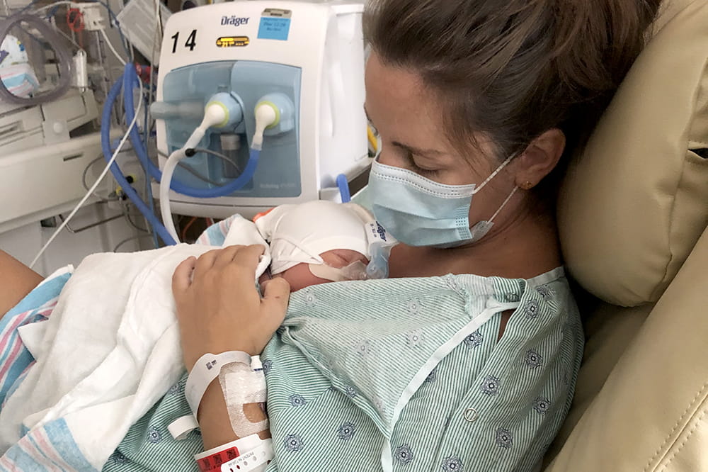 a woman in hospital gown and mask sits in a rocking chair holding a small baby wearing a CPAP device