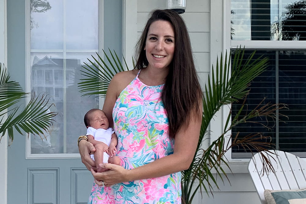 A smiling woman in a pastel floral sundress holds a tiny baby on a front porch